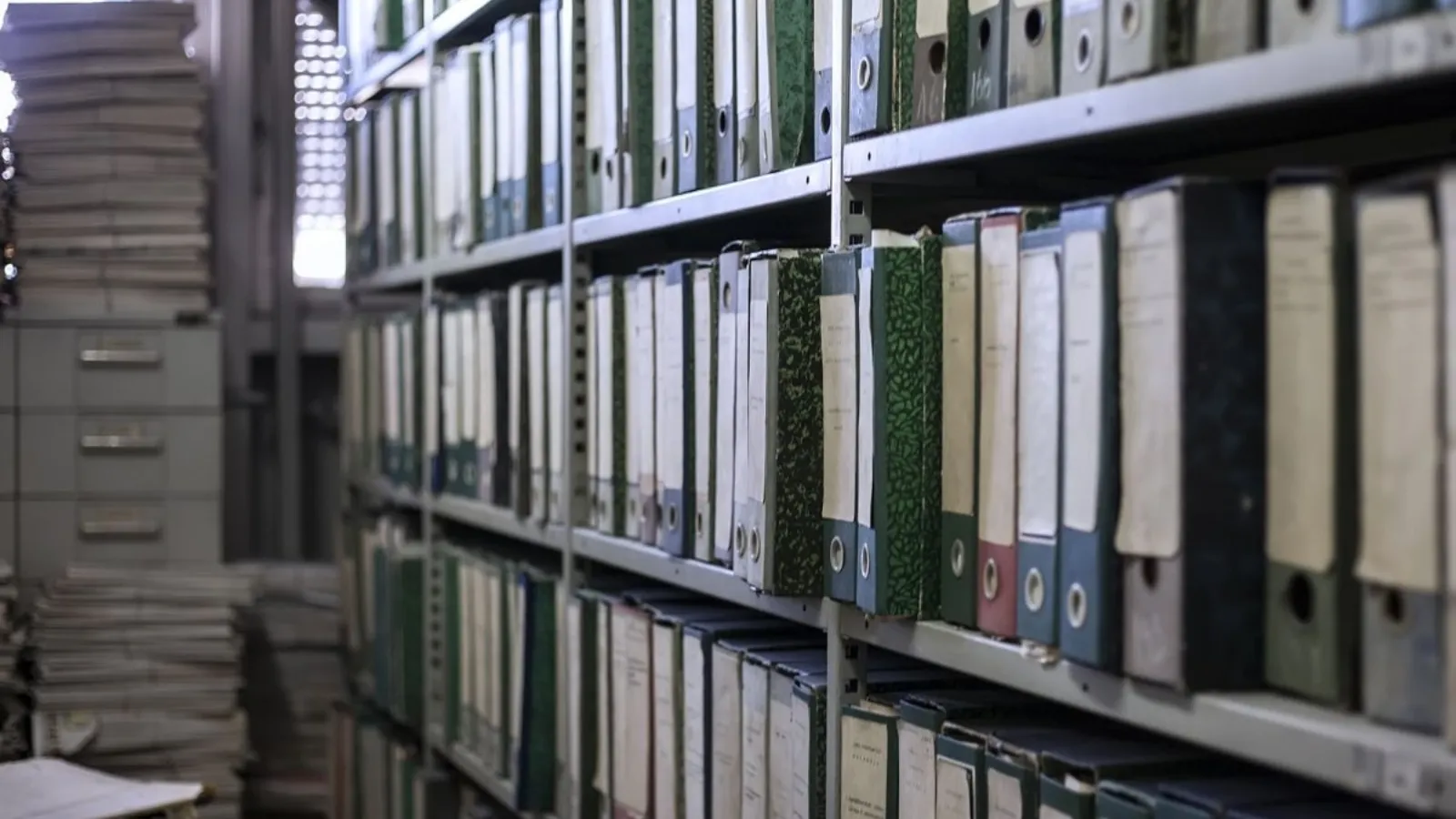 Shelves of ageing archive box files receding into a dim storeroom, with filing cabinets and stacked paper alongside.