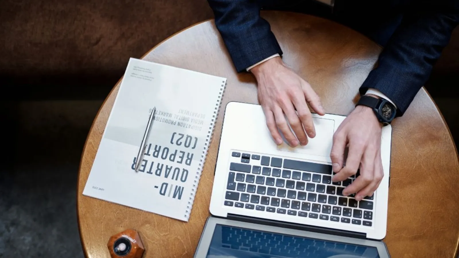 A top-down view of a man's hands typing on a laptop at a round wooden table, beside a printed report and a pen.