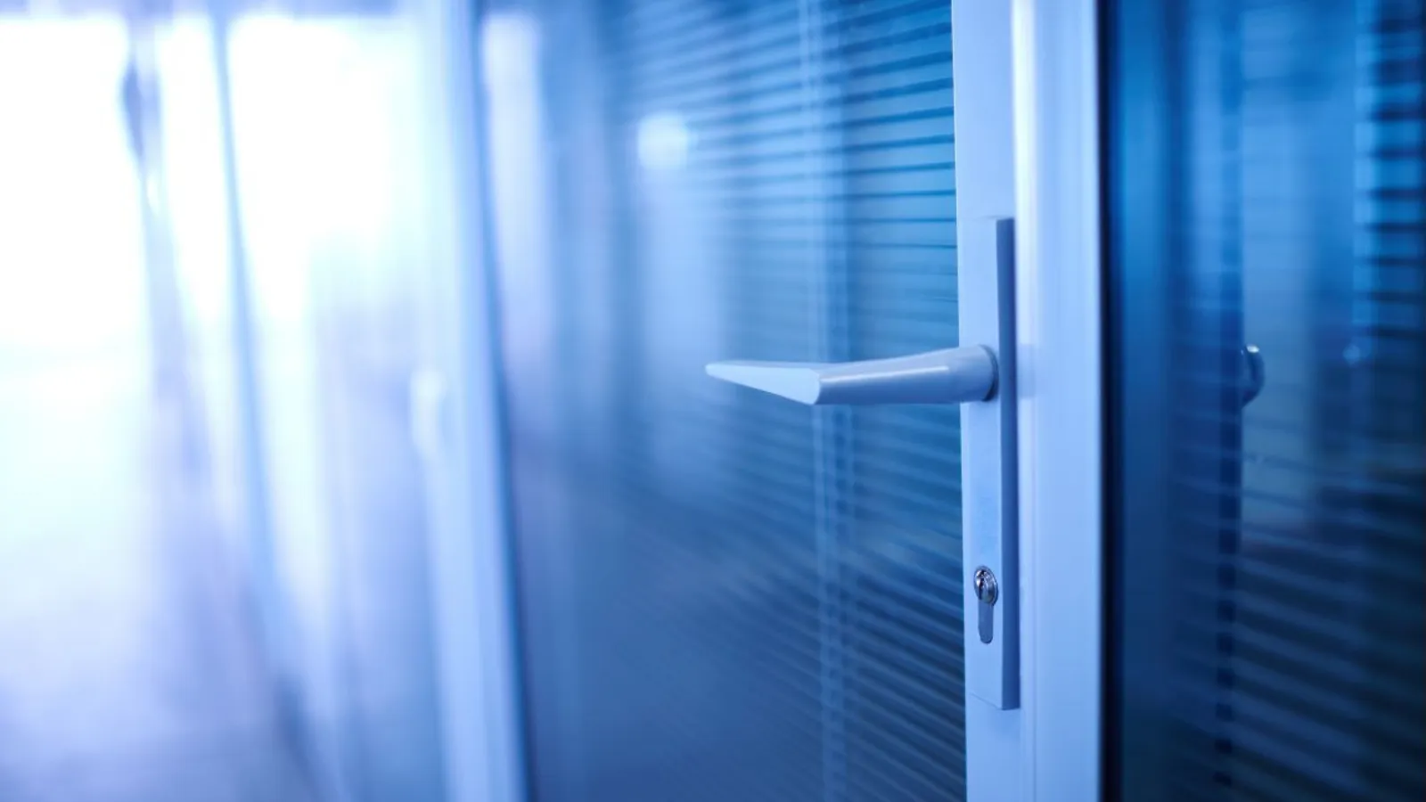 A closed glass office door in cool blue light, its handle in sharp focus and the corridor beyond blurred.