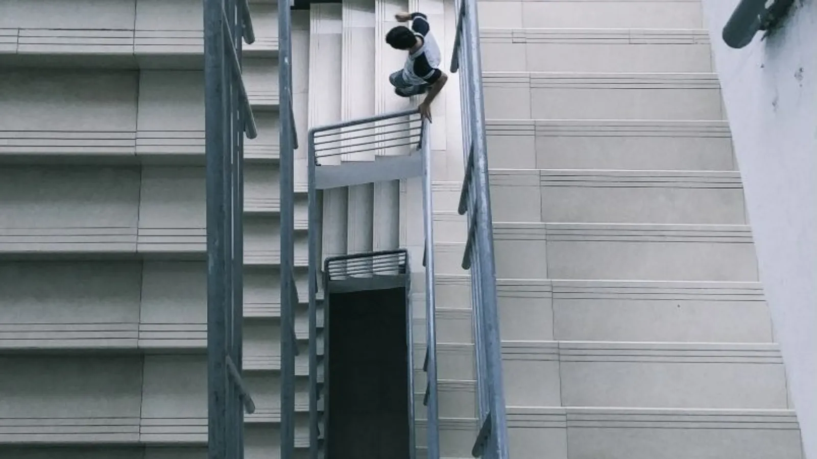 A person seen from above hurrying down a concrete stairwell, partly hidden behind the metal handrails of the flights between them.