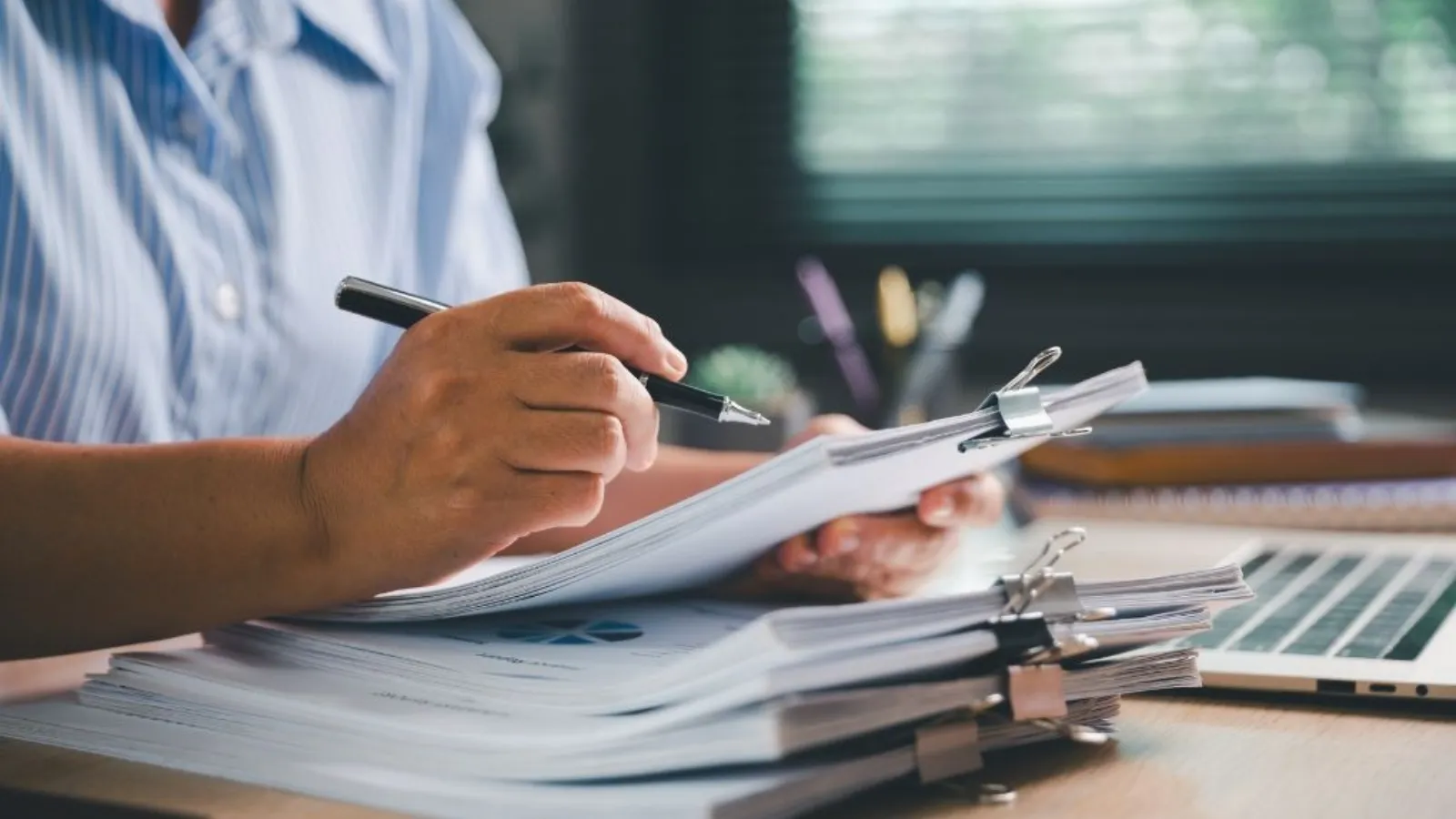 A hand holding a pen over a stack of clipped papers on a wooden desk, a laptop beside it.