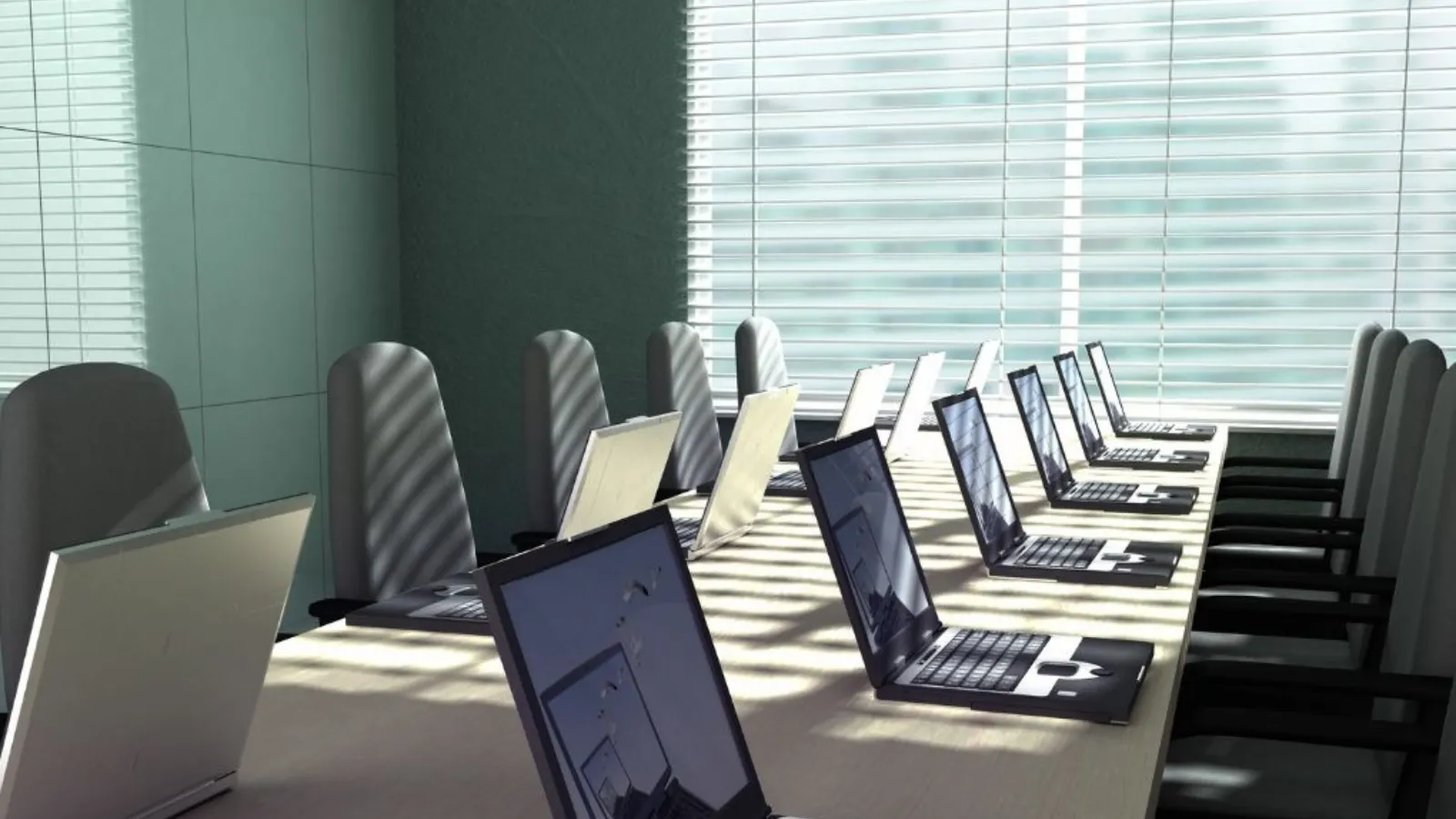 A long table in a bright meeting room with open laptops in a row and grey chairs, sunlight coming through the blinds.