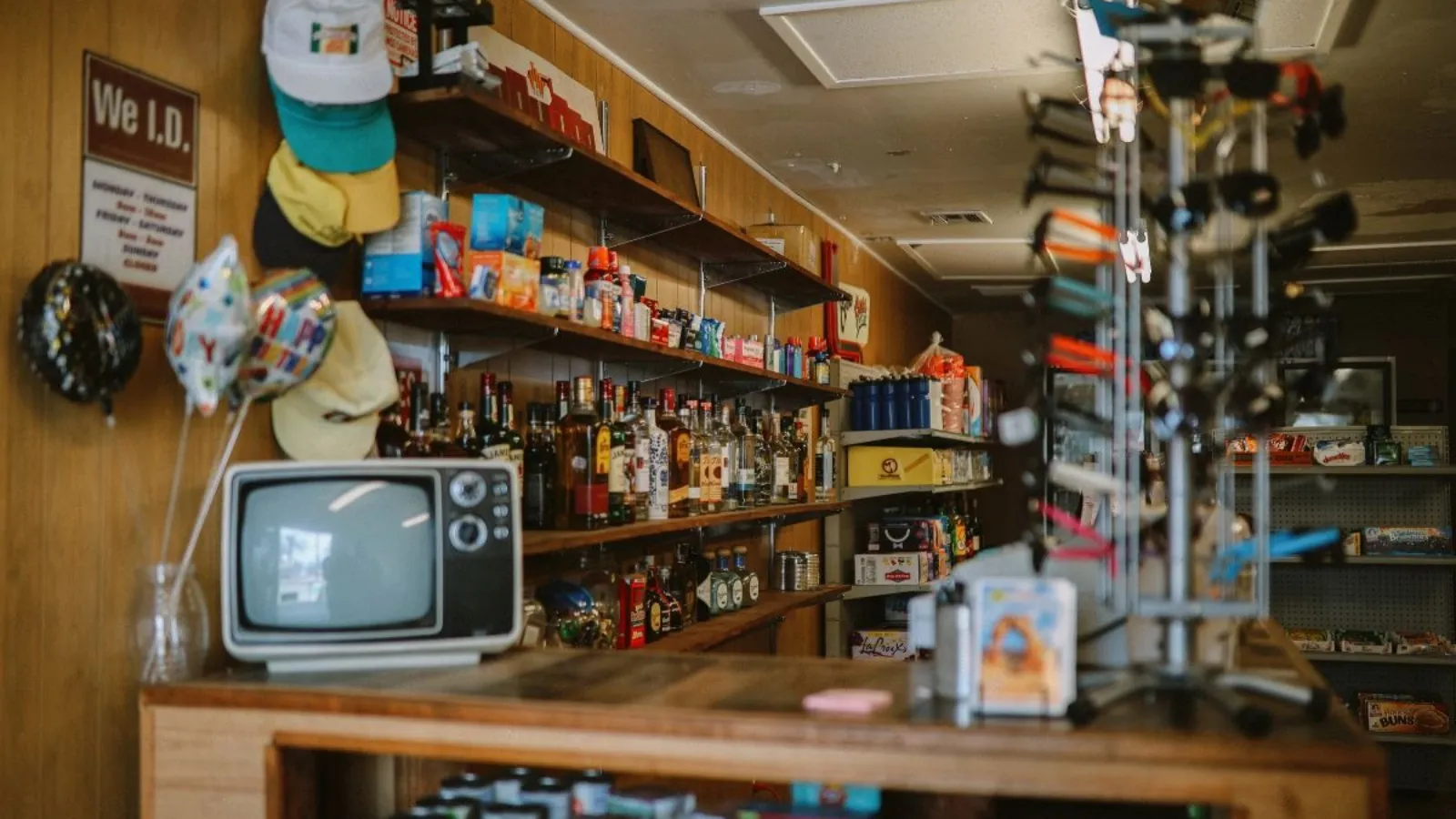 The inside of a small convenience shop, with wooden shelves of bottles and boxed goods above a counter, an old television and party balloons on the left.
