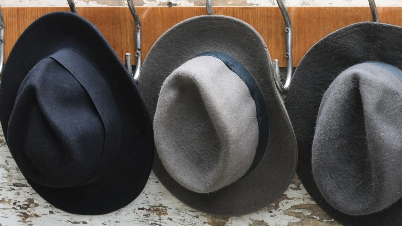 Three felt hats hanging side by side on hooks against a weathered white wall.