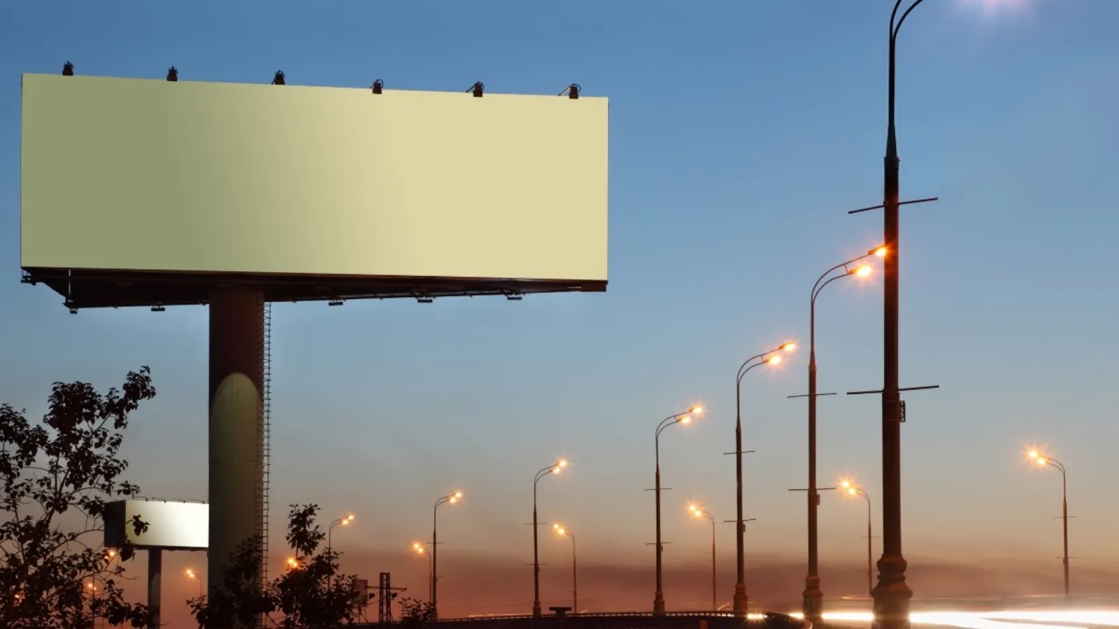 A blank roadside billboard at dusk, with a line of lit streetlights receding into the distance behind it.