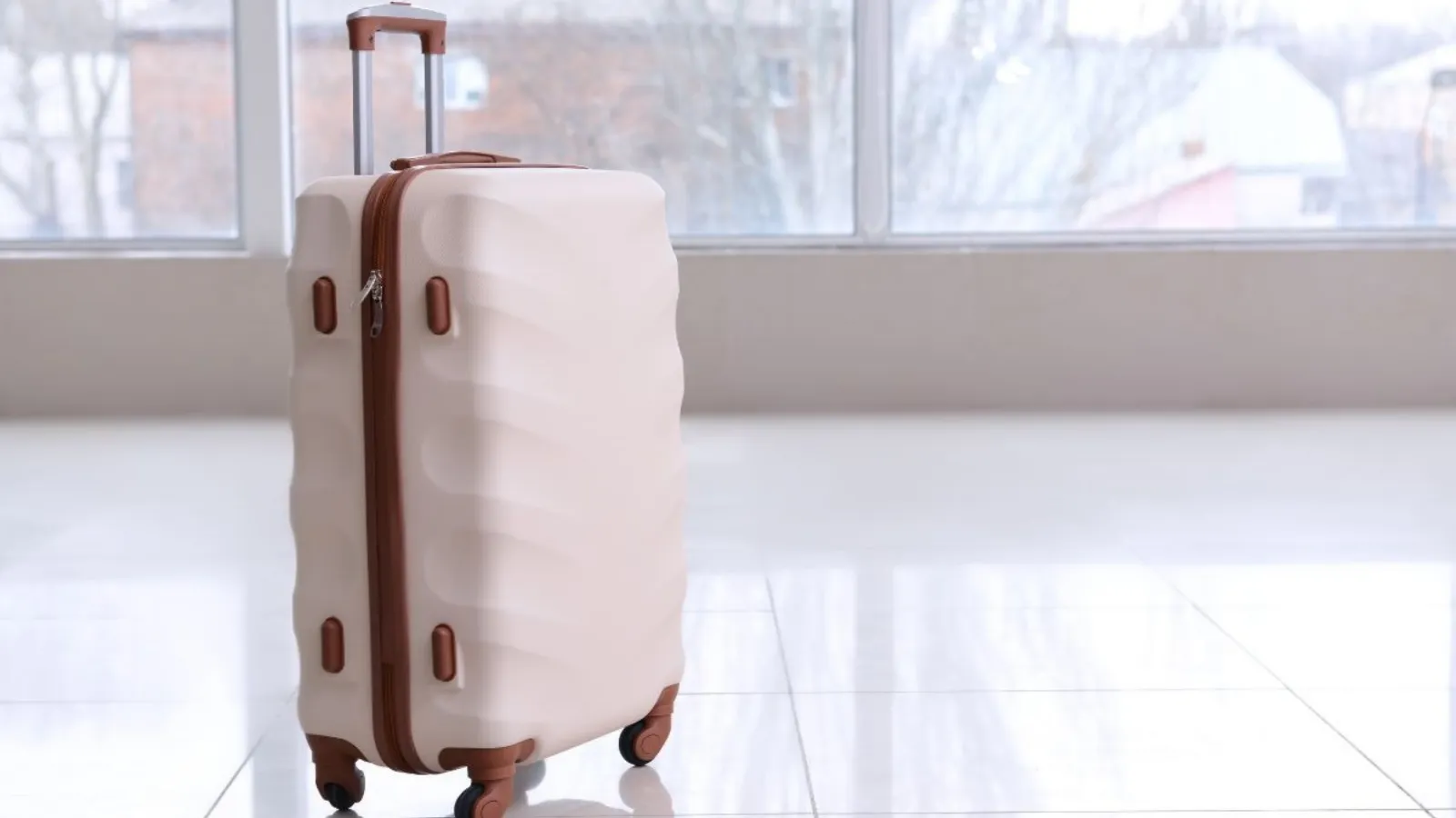 A cream suitcase standing on a shiny tiled floor in front of a large window.